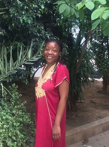 Una mujer sonriente con un vestido rojo con bordados dorados se encuentra frente a exuberantes plantas verdes, irradiando la tranquilidad que ofrece al dar los masajes holísticos Valencia.