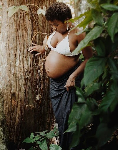 Una mujer embarazada con un top blanco y una falda azul está parada junto a un árbol en un bosque frondoso y holístico, tocándose suavemente el vientre y mirando pensativamente hacia abajo.