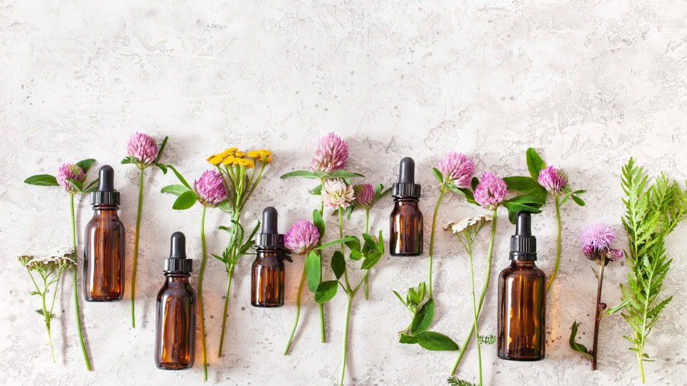 Brown dropper bottles are arranged with various wildflowers and green leaves on a light textured surface. The scene, featuring pink clover and yellow blossoms, captures a natural and earthy aesthetic reminiscent of a tranquil morning run in the lush landscapes surrounding Valencia.